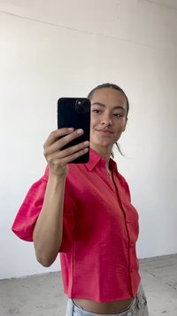 Cămașă roz aprins cu mânecă scurtă și butoni, dintr-un material texturat, purtată cu blugi albaștri deschiși. Subiectul ține un smartphone în mâna dreaptă.