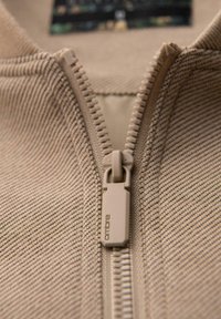 Beżowy zamek błyskawiczny z fakturowanej tkaniny, z wyraźnym suwakiem prostokątnym oraz metką marki w tle.