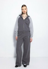 Młoda kobieta stoi przy białej ścianie, ubrana w szary dres z zamkiem błyskawicznym i jaśniejszymi rękawami oraz pasujące szerokie spodnie i czarne buty.