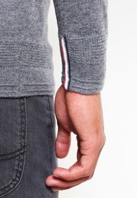 Grauer Strickpullover aus strukturiertem Material, mit einem rot, weiß und blau gestreiften Akzent am Bündchen. Die Hand liegt auf einer dunkelgrauen Hose.