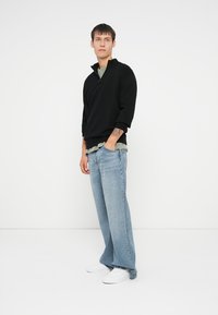 Black pullover with a zip collar, layered over a green shirt. Light blue wide-leg jeans and white sneakers complete the outfit.