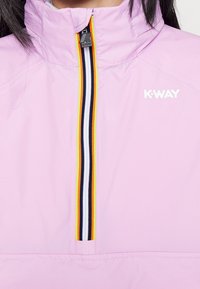 Světle růžová bunda K-Way s vysokým límcem, se zipem a černými, bílými, žlutými a oranžovými pruhy, a logem na hrudi.
