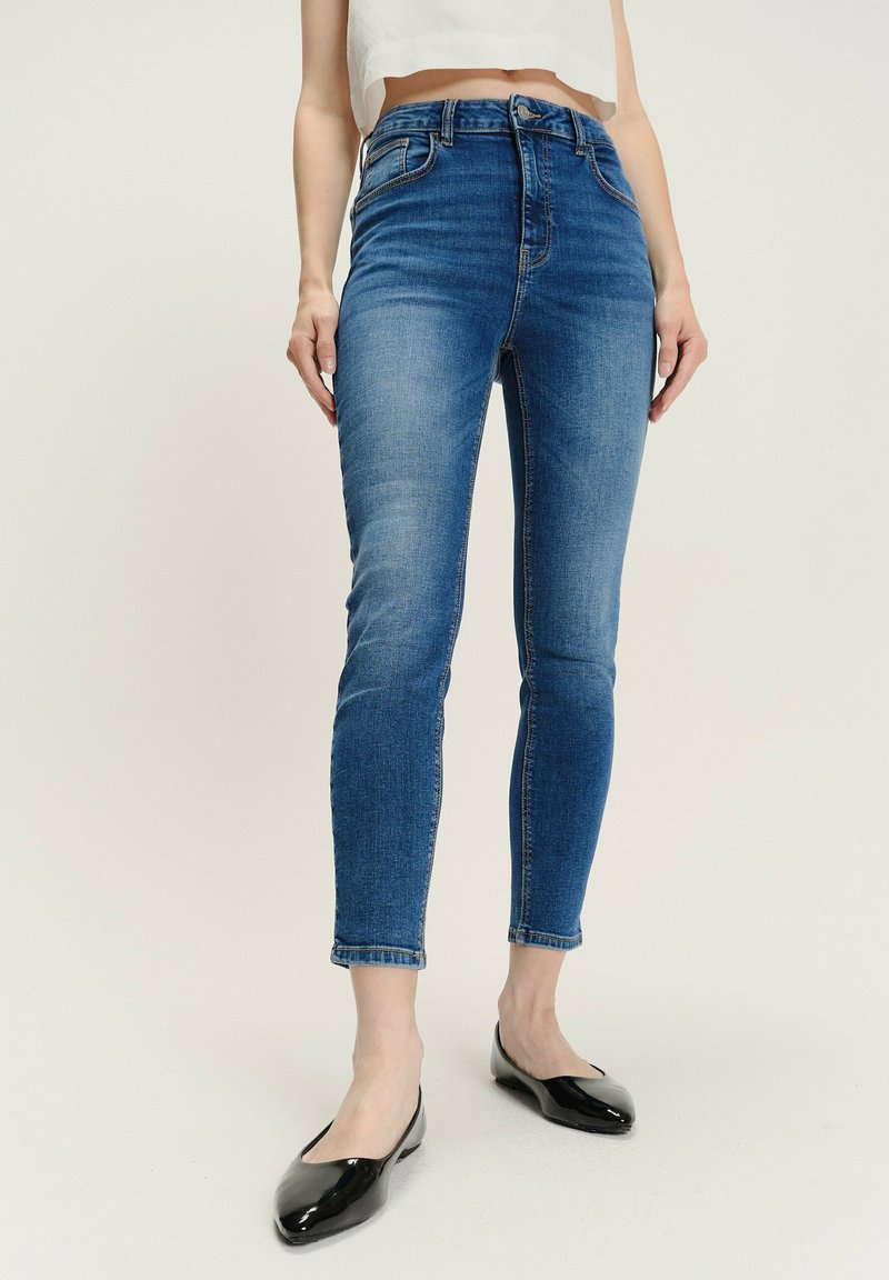 Kobieta w niebieskich, slim-fit, wysokich jeansach oraz czarnych, lakierowanych balerinach, stoi na tle jasnym.