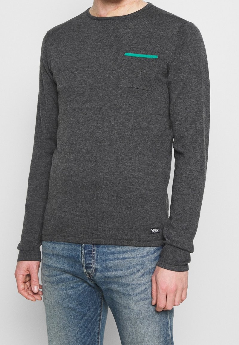 Homme portant un pull gris foncé à manches longues avec une petite poche poitrine accentuée de vert et un jean bleu délavé, debout devant un fond blanc.