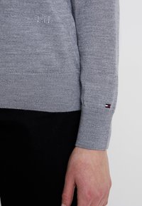 Harmaa neule, jossa on ribattu helma ja hihansuut, ja joka sisältää monimutkaisen neuletekstuuri. Pieni logo-tila punaisena, valkoisena ja sinisenä hihassa.