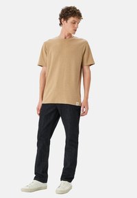 T-shirt beige a maniche corte con collo tondo, abbinato a jeans blu scuro e sneaker bianche, con un piccolo etichetta sull'orlo della t-shirt.