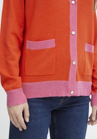Orange kofta med rosa ribbade manschetter och fåll. Har två rosa fickor och vita knappar. Mjuk textur, avslappnad passform, ärmar i medellängd.