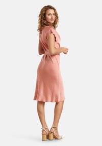 Rosa knielanges Kleid aus leichtem Stoff mit kurzen Ärmeln und einem taillierten Gürtel. Ergänzt durch beigefarbene Sandalen mit Blockabsatz.
