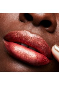 Metaalachtige rode lipstick op volle lippen, die de textuur en glans accentueert tegen een donkere huidtint; een vinger raakt voorzichtig een lip aan voor extra nadruk.