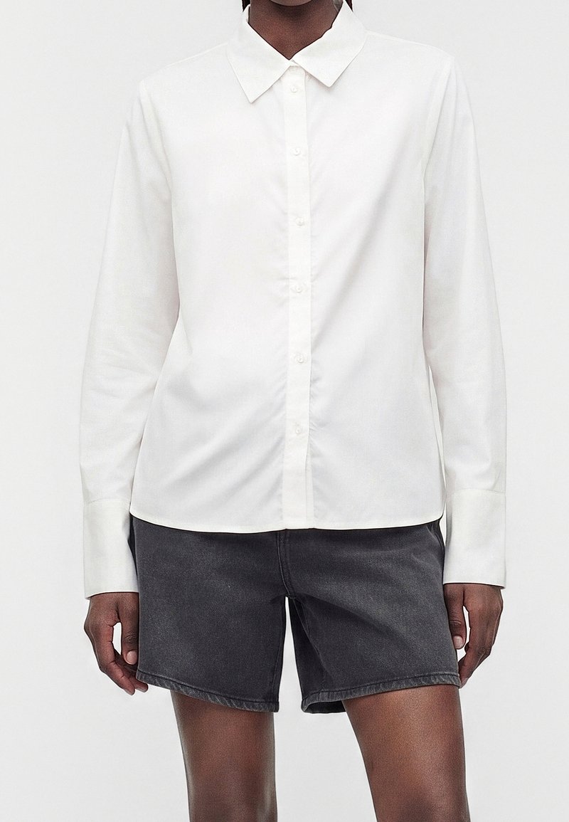 Personne portant une chemise blanche à manches longues boutonnée et un short en jean gris foncé sur un fond clair uni.