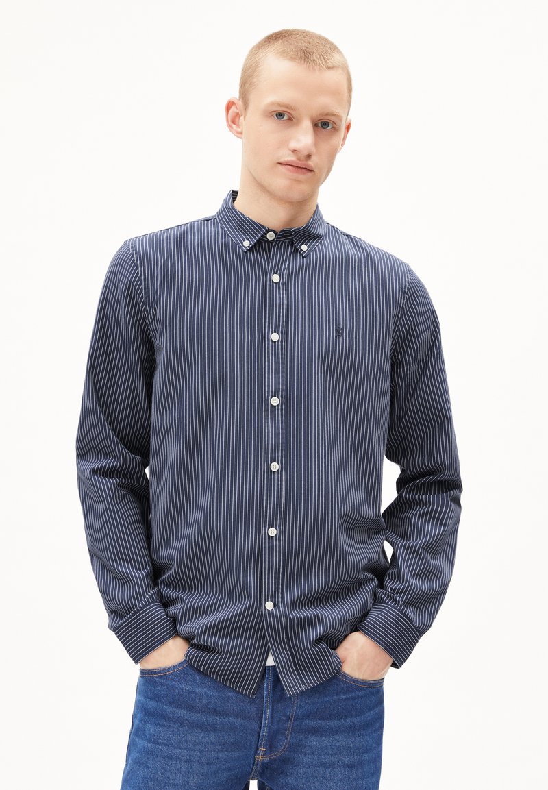 Navy gestreiftes Button-Up-Hem mit langen Ärmeln und Kragen; enthält weiße Knöpfe und ein kleines Logo auf der Brust. Getragen mit blauen Jeans.