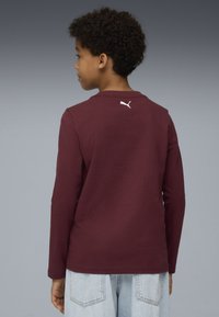 Bordeauxrotes Langarmshirt mit Rundhalsausschnitt. Auf der Rückseite befindet sich ein weißes Puma-Logo. Kombiniert mit hellblauen Jeansshorts.