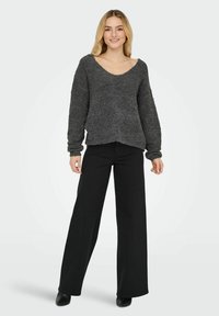 Pilkas tekstūruotas megztinis su plačiu apykaklė, ilgomis rankovėmis, derinamas su juodomis plačiomis kelnių ir juodais batais, stovintis ant vientiso fono.