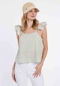 Top sin mangas a rayas con acentos de hombros volantes, en colores verde claro y blanco. Combinado con pantalones blancos. Lleva una gorra de paja beige.