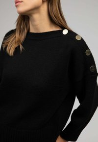 Pull en maille noir avec un décolleté bateau, décoré de boutons en métal doré sur l'épaule gauche et de poignets côtelés. Texture douce et coupe décontractée.