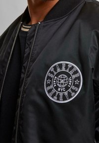 Veste aviateur noire avec une texture lisse et brillante. Elle présente un logo rond brodé en blanc sur la poitrine avec l'inscription "STARTER" et un design de globe.