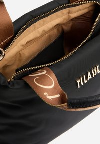 Borsa nera in nylon con una zip dorata, foderata con tessuto marrone. Presenta un logo sul lato e una tracolla color cuoio con un motivo decorativo.