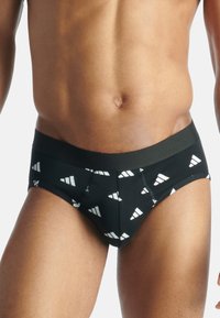 Mustat alushousut, jotka on valmistettu joustavasta kankaasta ja joissa on valkoinen kolmiomainen kuvio. Vyötärönauha on leveä ja yhtenäinen musta, ja saumat ovat näkyvillä.