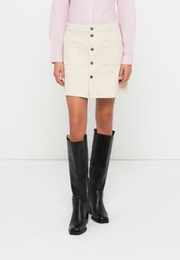 Person som har på seg et beige knapp-front skjørt, en rosa stripete langermet skjorte, og svarte knelange høye hæler som står på et hvitt gulv.