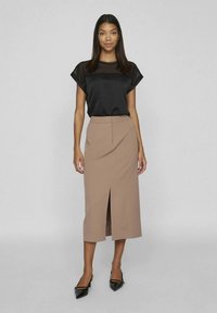 Donna che indossa una camicetta nera a maniche corte e una gonna midi beige con una spacco sul davanti, abbinata a scarpe con tacco nere a punta, in piedi su uno sfondo grigio.