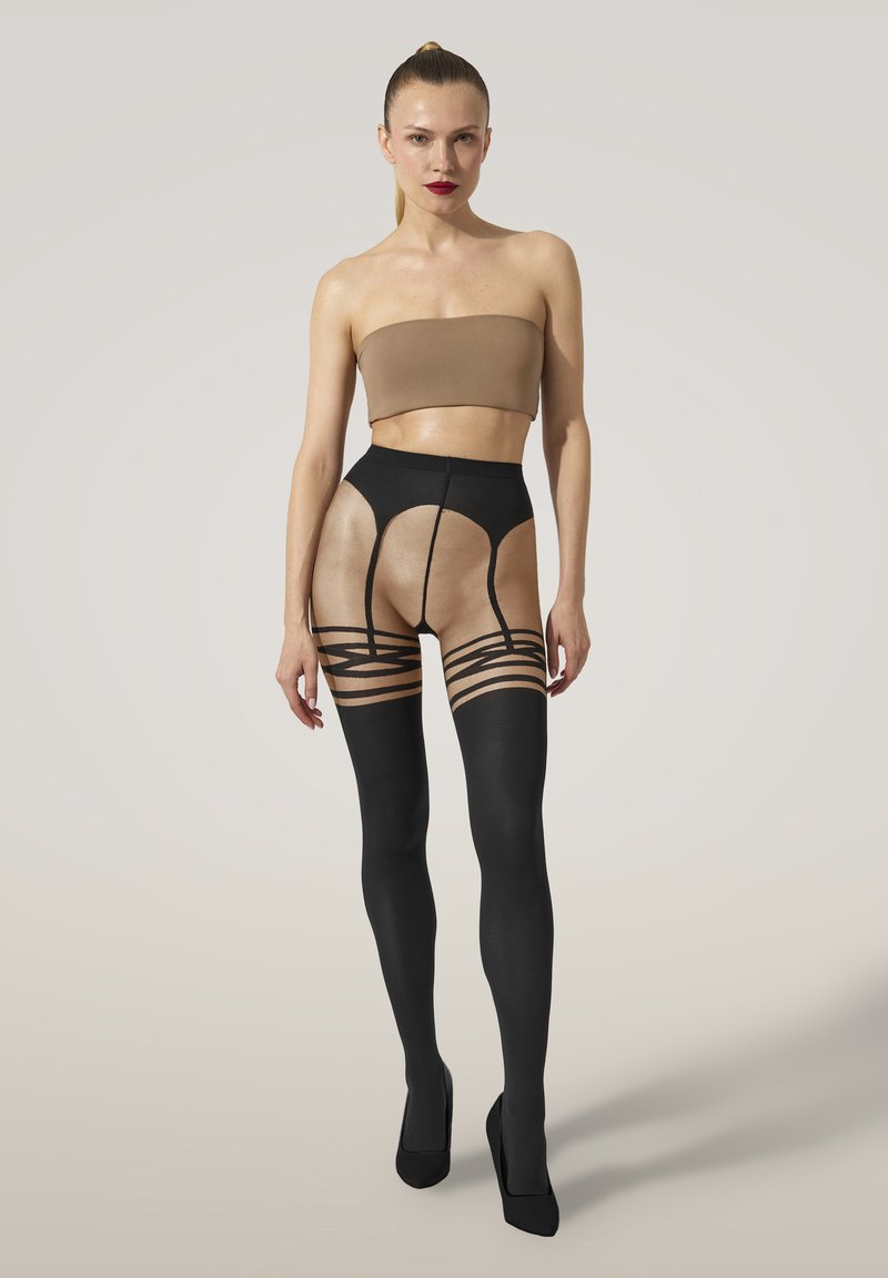 Kvinna iklädd högmidjade svarta mönstrade genomskinliga strumpbyxor, brun axelbandslös crop top och svarta högklackade skor, stående mot en ljus bakgrund.