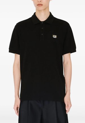 Person iført en sort kortærmet poloshirt med tre knapper og et lille flerfarvet broderet logo på venstre bryst.