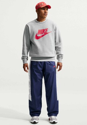 Jonge man draagt een grijze Nike trui, marineblauwe trainingsbroek, witte sneakers en een rode pet met vlammenpatroon, kijkt opzij.