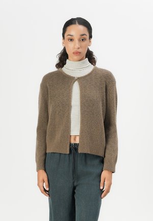 Brun strikket cardigan med rund hals, åpen frem, og uten knapper. Brukt over en stripete høyhalset genser, med lange ermer og en kort design.