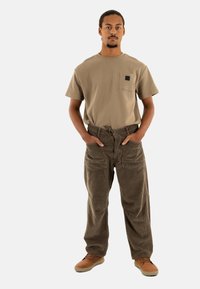 Camiseta beige de manga corta con bolsillo; combinada con pantalones de pana marrones que tienen múltiples bolsillos y un corte relajado. Zapatos de color sólido.