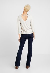 Top beige clair à manches longues avec un détail en forme de trou de serrure à l'arrière, associé à un jean évasé bleu foncé. Tissu texturé avec une coupe décontractée.