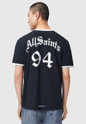 Man wearing black T-shirt with white trim, "AllSaints 94" text on back, paired with blue jeans, standing against plain gray background.