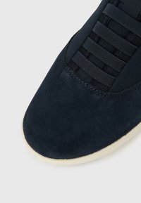 Κοντινή όψη στο μπροστινό μέρος ενός σκούρου μπλε σουεντ αθλητικού παπουτσιού με  μαύρο δίχτυ και κορδόνια, καθώς και λευκή σόλα σε φωτεινό φόντο.