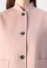 Hellrosa Wollmischungsmantel mit hohem Kragen, ausgestattet mit vier Knöpfen und zwei Fronttaschen. Weiche Textur und minimalistisches Design.