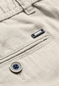 Gros plan d'un pantalon en tissu beige montrant une poche avec un bouton blanc et une petite étiquette métallique de la marque Bugatti au-dessus de la poche.