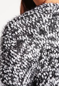 Tjockstickad tröja i svart och vitt med ett strukturerat mönster, som har mjuka, fluffiga fibrer och lös passform. Accenter av glansiga trådar syns.