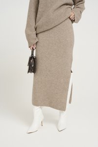 Beiges, geripptes Strickoberteil und Rockset mit seitlichen Schlitzen, kombiniert mit weißen Stiletto-Stiefeln. Hält eine kleine schwarze Handtasche.