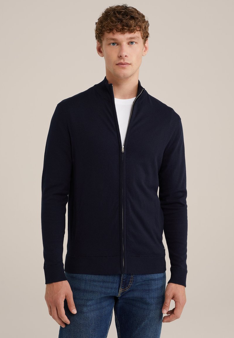 Marinblå zip-up cardigan i ett slätt stickat material, med hög krage och ribbade muddar. Bärs över en vit skjorta, tillsammans med blå jeans.