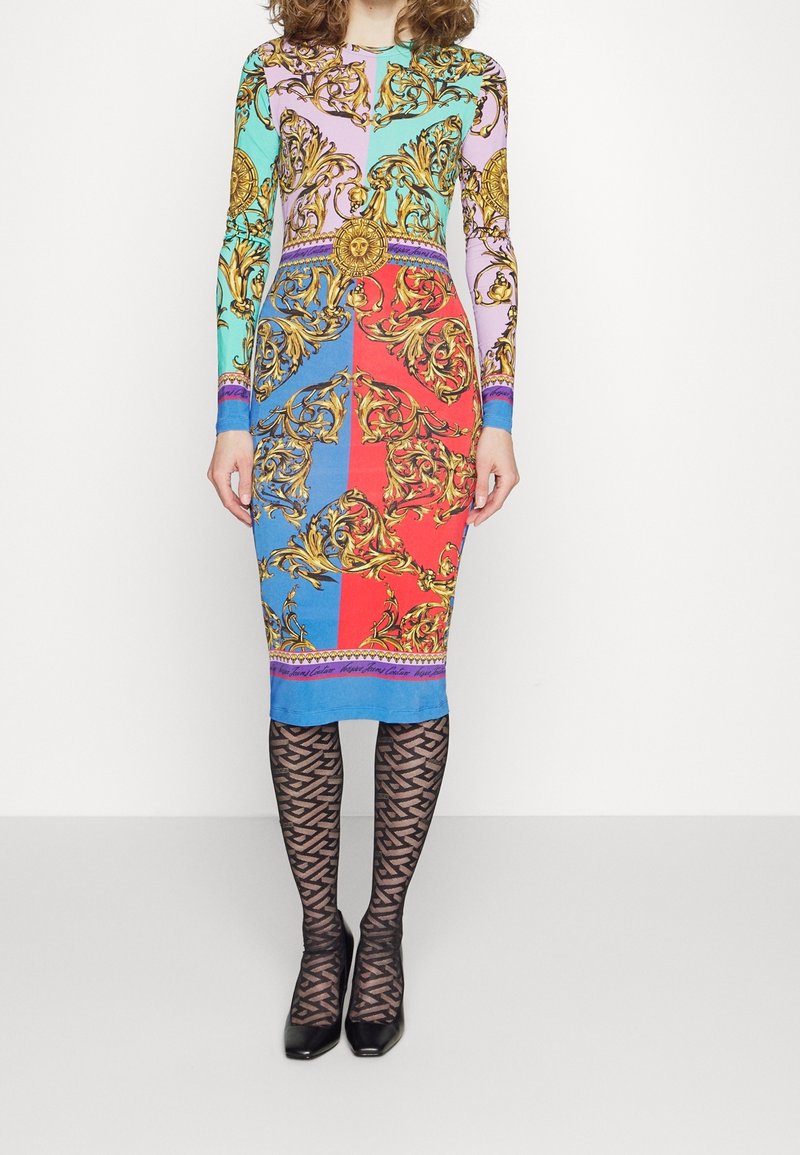 Femme portant une robe midi à blocs de couleurs avec des motifs baroques dorés, associée à des collants noirs à motifs et des talons noirs à bout pointu.