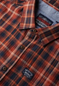 Red and navy plaid shirt made of fabric with a textured finish. Features a button-down collar and a branded patch near the bottom.