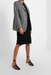 Blaser en tweed gris avec revers cranté, fermeture à bouton unique, deux poches avant, porté sur une robe noire, associé à des chaussures noires à lanières.