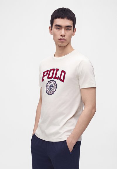 Νέος άνδρας που φοράει λευκό t-shirt Polo Ralph Lauren με κόκκινο λογότυπο και σκούρο μπλε παντελόνι, στέκεται με τα χέρια στις τσέπες μπροστά σε απλό φόντο.