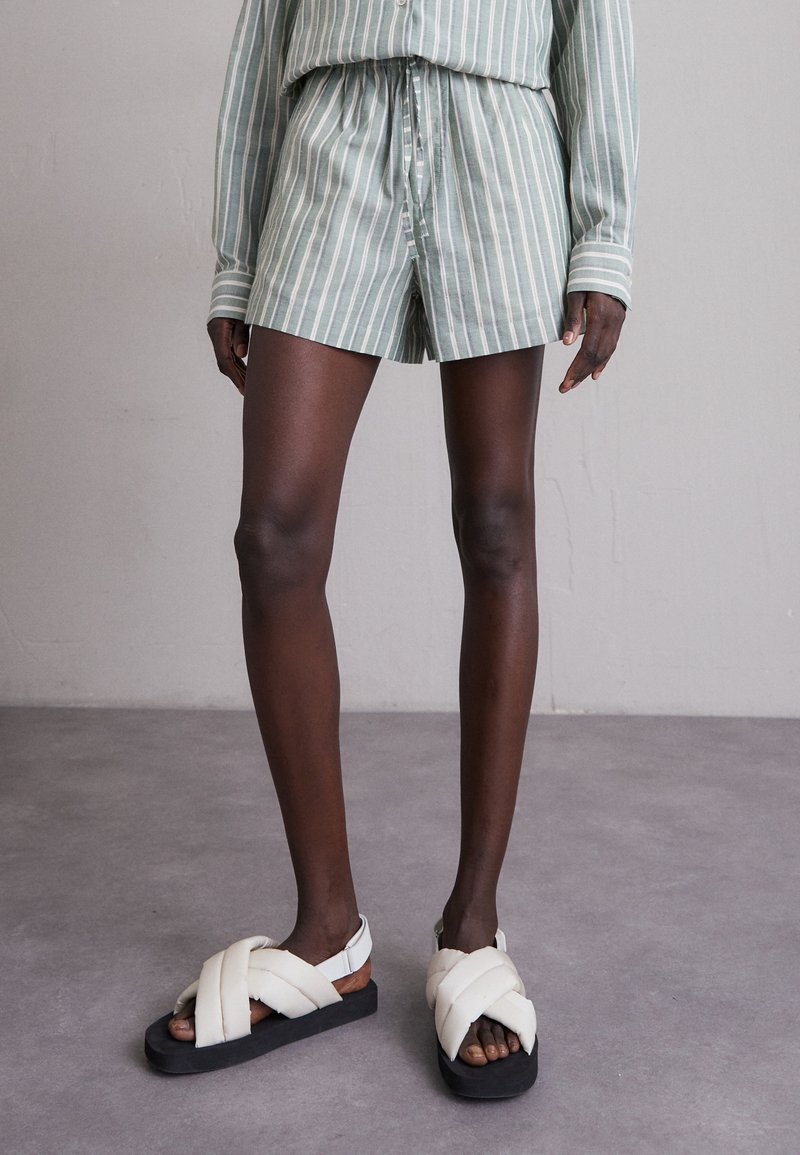 Personne portant un short à rayures verticales vertes et blanches ainsi qu'une chemise longue assortie, avec des sandales blanches rembourrées sur un sol gris.