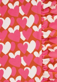 Textilný vzor s ružovými a bielymi srdiečkami na hnedom pozadí, so zúbkovaným okrajom na pravej strane.