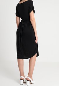 Mustapuku, jossa on lyhyet hihat, vyötärösolki ja kaareva helma. Yhdistetty valkoisiin paksupohjaisiin sandaaleihin. Pehmeä, sileä kangas.