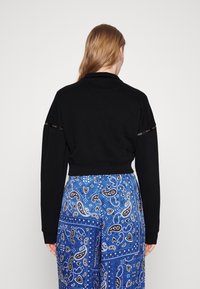 Sudadera negra corta con mangas largas y dobladillo acanalado, con acento de logo en las mangas. Combinada con pantalones azules de patrón paisley.