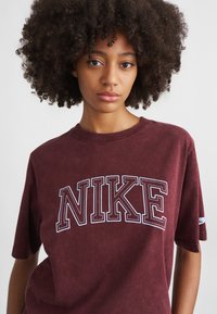 Kāpurpura kokvilnas T-krekls ar lielu balto izšūto "NIKE" logo. Tam ir apaļa kakla izgriezums un īsas piedurknes ar mīkstu, teksturētu apdari.