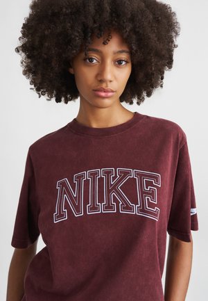 Kāpurpura kokvilnas T-krekls ar lielu balto izšūto "NIKE" logo. Tam ir apaļa kakla izgriezums un īsas piedurknes ar mīkstu, teksturētu apdari.