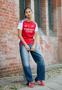 Červený a bílý dres Adidas s logem "Emirates Fly Better", doplněný volnými modrými denimovými džínami a červenými teniskami. Pozadí tvoří cihlová zeď.