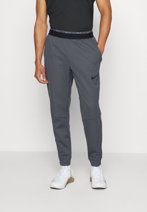 Homme portant un pantalon de jogging Nike gris foncé avec un logo noir, un t-shirt noir et des chaussures de sport blanches sur un fond clair uni.
