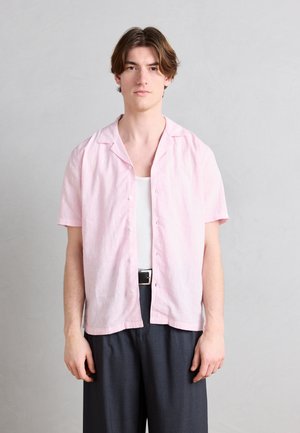 Giovane uomo con capelli castano medi, indossa una camicia rosa chiaro a maniche corte abbottonata sopra una canottiera bianca e pantaloni scuri.